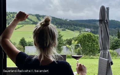 Frau steht mit einem Glas Wein vor einem Fenster, welches die grüne Landschaft im Sauerland zeigt.