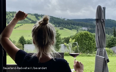 Vrouw staat met een glas wijn voor een raam met het groene landschap van het Sauerland.