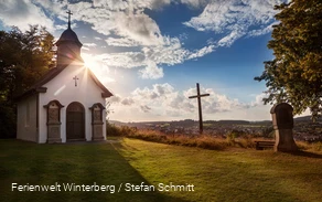 Eine Kapelle neben einem Kreuz und einer Kreuzwegstation mit blauem Himmel und der Sonne im Hintergrund.